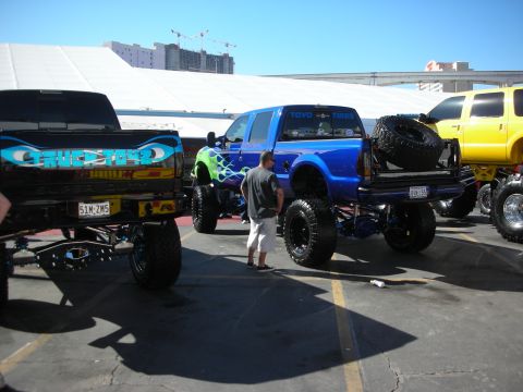 1998 Ford Crown Victoria - Lineup of brightly painted lifted pickups, including a flamed blue truck, in an outdoor show lot beside a domed venue.