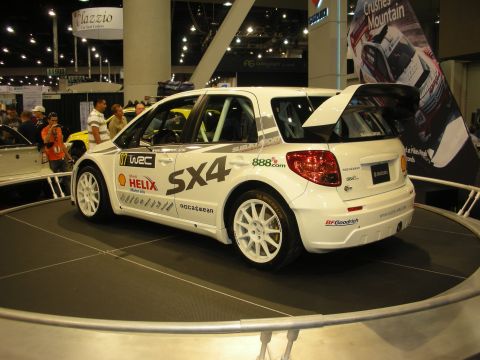 1998 Ford Crown Victoria - white Suzuki SX4 rally car liveried with WRC and sponsor decals sits on a rotating display stand at an expo.