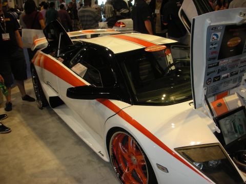 1998 Ford Crown Victoria - white supercar with an orange center stripe sits with its scissor door raised beside a vendor display at a show.