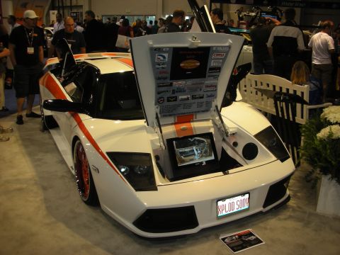 1998 Ford Crown Victoria - white Lamborghini Murcielago sits with hood and scissor doors raised, displaying a sponsor-covered panel at an expo.