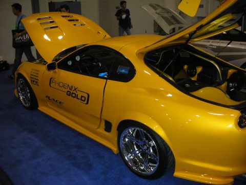 1998 Ford Crown Victoria - yellow sports car with a vented hood, chrome multi-spoke wheels and Phoenix Gold audio decals on display indoors.