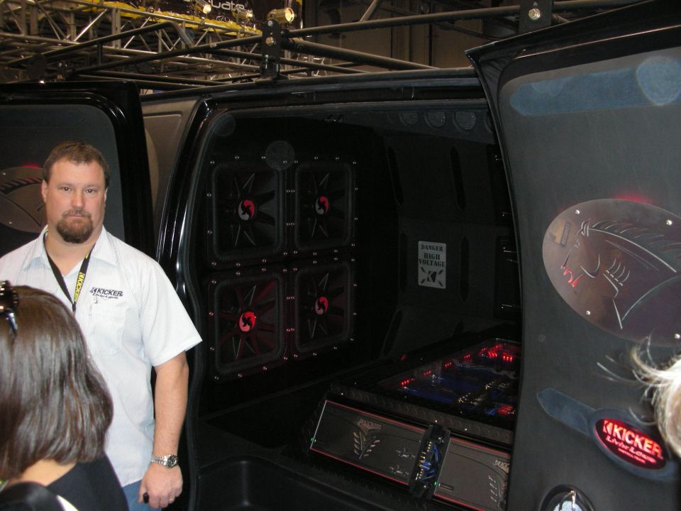 Man beside black van with four red-lit amplifiers and Kicker audio display