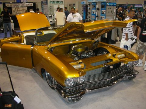 1998 Ford Crown Victoria - gold custom classic car sits with hood raised showing a chromed engine, surrounded by attendees at an expo.