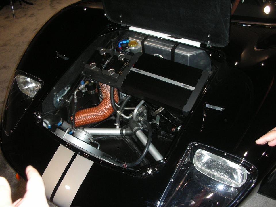 Open front trunk of a black sports car showing fuel tank, plumbing and a coiled copper duct