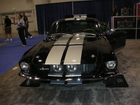 1998 Ford Crown Victoria - black fastback Mustang with twin silver racing stripes sits on display at an indoor car show with onlookers nearby.