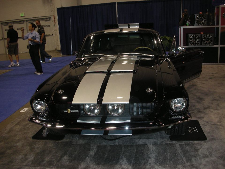 Front of a black fastback Mustang with silver racing stripes at an indoor car show