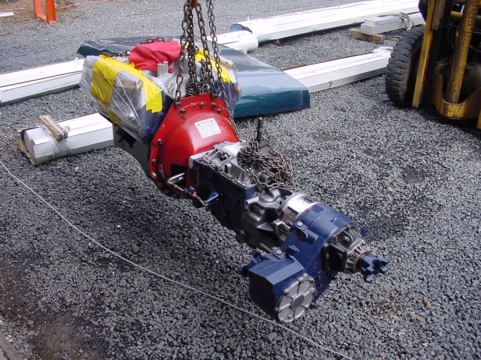 Transmission and transfer case assembly suspended by chains, with a forklift visible in the background