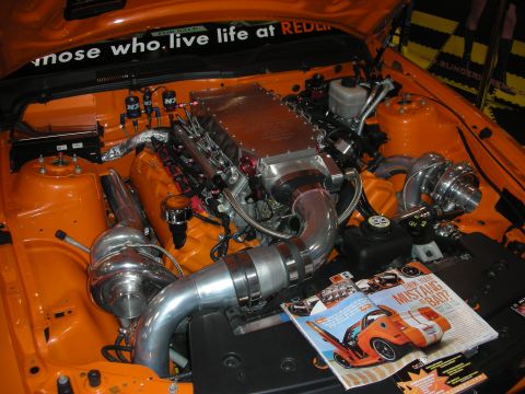 1998 Ford Crown Victoria - orange engine bay holds a twin-turbo V8 with polished piping and braided lines, a magazine propped in front.