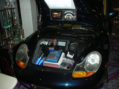 1998 Ford Crown Victoria - front trunk of a dark Porsche Boxster is filled with audio equipment, a mixer, screen and a game controller.