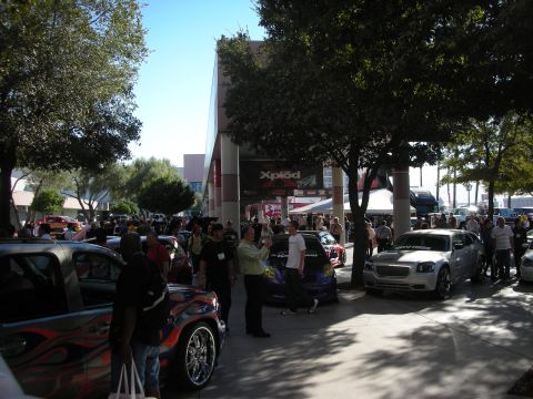 1998 Ford Crown Victoria - Custom cars and a crowd fill an outdoor show plaza beneath trees, with an Xplod banner on the building behind.