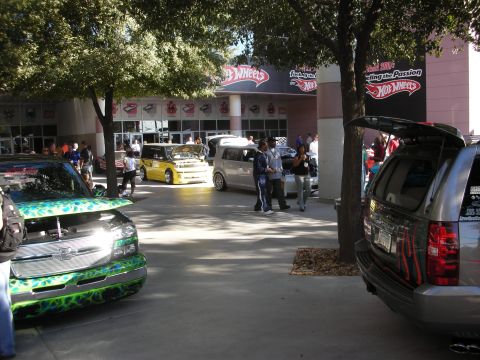1998 Ford Crown Victoria - Custom cars including a green-flamed truck and a black SUV sit outside a Hot Wheels storefront at a show.