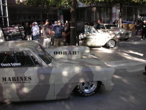 1998 Ford Crown Victoria - Two camouflage-painted muscle cars with Marines and OORAH! lettering sit on display outdoors among onlookers.