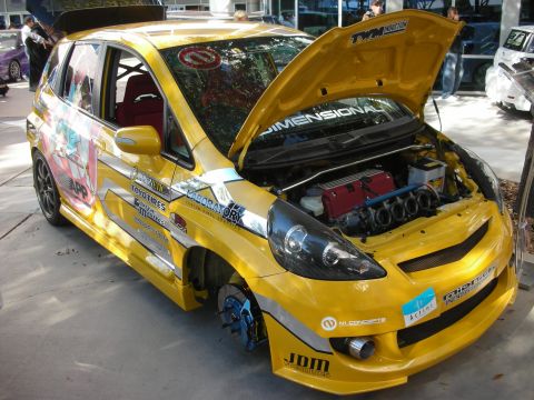 1998 Ford Crown Victoria - yellow hatchback covered in anime graphics and sponsor decals sits with hood open exposing an engine, one wheel removed.