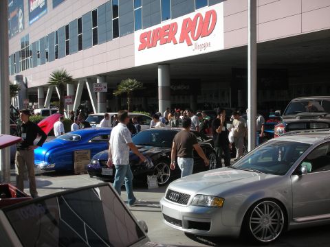 1998 Ford Crown Victoria - crowd browses custom cars beneath a Super Rod Magazine sign, with a silver Audi sedan parked in the foreground.