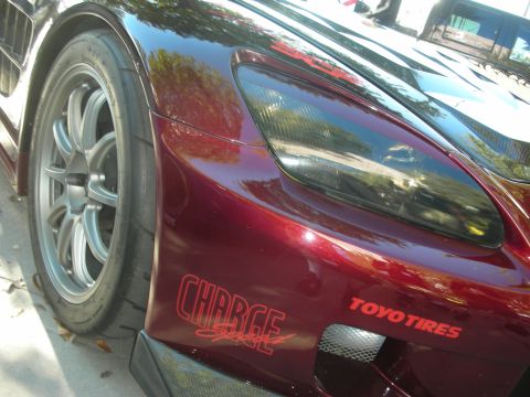 1998 Ford Crown Victoria - close-up of a dark red sports car shows its front fender, headlight, silver wheel and Charge and Toyo Tires decals.