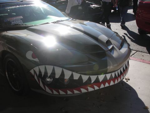 1998 Ford Crown Victoria - front of a coupe painted with military shark-mouth teeth and eyes, parked outdoors among other cars at an event.