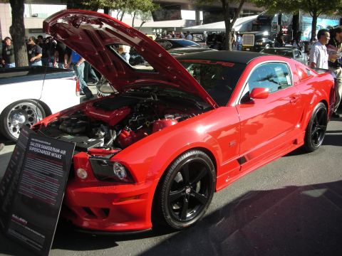 1998 Ford Crown Victoria - red Saleen-badged Ford Mustang coupe displayed with its hood open and an info placard, parked on red carpet at a show.