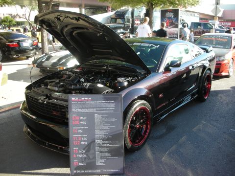 1998 Ford Crown Victoria - black Saleen Mustang with red-lipped wheels sits hood-up beside a spec placard listing 530 WHP at an outdoor show.