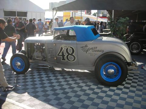 1998 Ford Crown Victoria - silver open-wheel hot rod roadster with blue accents and Honest Charley Since 48 lettering displayed on a checkered floor indoors.