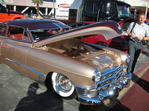 1998 Ford Crown Victoria - gold lowered custom Cadillac fastback with whitewall tires and hood open, displayed at an outdoor car show with a man nearby.