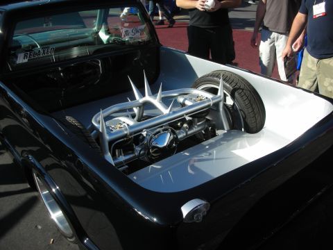 1998 Ford Crown Victoria - black custom truck bed opened to reveal a chrome V engine with dramatic spiked valve covers and a spare tire inside.