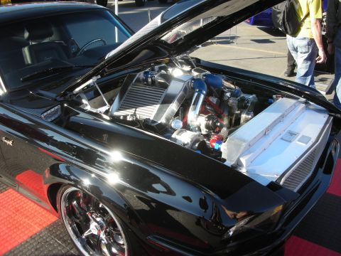 1998 Ford Crown Victoria - black muscle car shown hood-up on chrome wheels, revealing a turbocharged engine setup, parked on red flooring at an event.