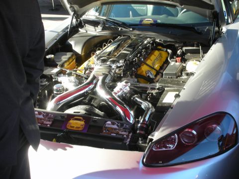 1998 Ford Crown Victoria - silver coupe with hood open showing a polished engine with chrome long-tube headers and yellow valve covers at an outdoor show.