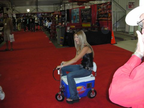 1998 Ford Crown Victoria - woman in a leather vest rides a motorized cooler-scooter down a red carpet aisle at an indoor automotive trade show.