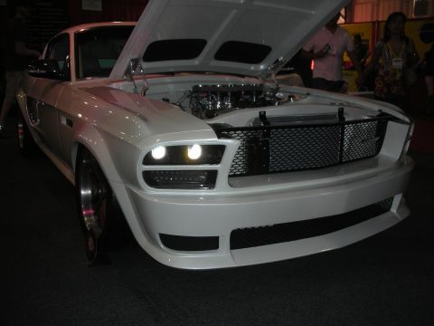 1998 Ford Crown Victoria - White custom Mustang fastback with open hood and aggressive front fascia displayed at an indoor automotive show.