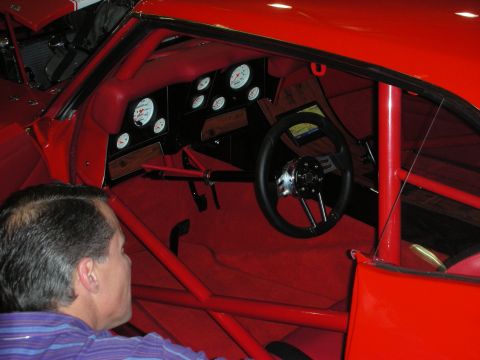 1998 Ford Crown Victoria - Red race-car interior with full roll cage, white-faced gauges, and a small steering wheel, seen over a spectator's shoulder.