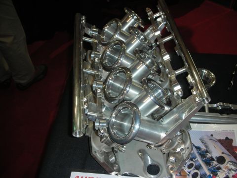 1998 Ford Crown Victoria - Polished aluminum V8 intake manifold with individual velocity stacks displayed on a stand at a trade show.