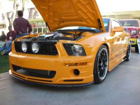 1998 Ford Crown Victoria - Bright orange Ford Mustang coupe displayed with its hood open at an outdoor car show, fitted with a custom mesh grille and black multi-spok…