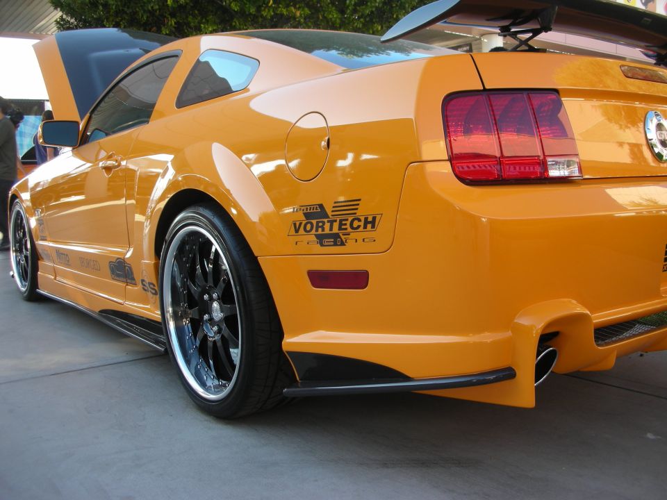 Rear three-quarter of an orange Mustang with Vortech decal and chrome wheels