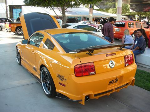 1998 Ford Crown Victoria - Orange Ford Mustang shown rear three-quarter with hood up and rear wing, parked among other cars at an indoor-outdoor show.