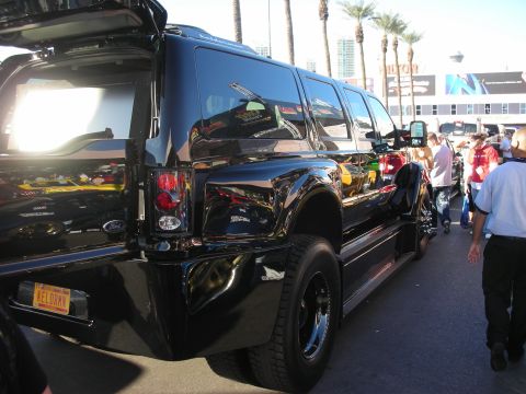 1998 Ford Crown Victoria - Black stretched Ford Excursion-style SUV limousine with extra side windows parked along a street near palm trees.
