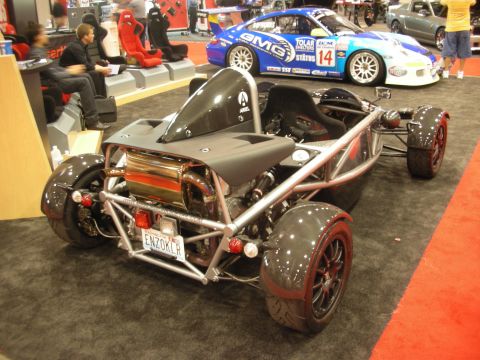 1998 Ford Crown Victoria - Gray skeletal exoskeleton track car shown rear three-quarter with a blue and white race car parked behind at an indoor expo.