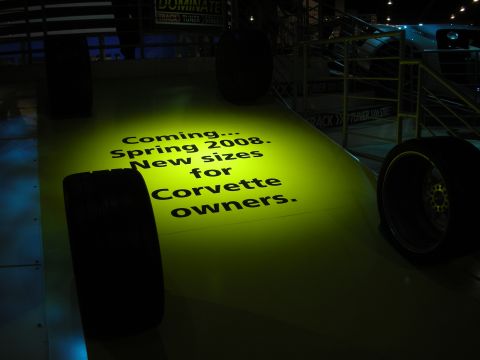 1998 Ford Crown Victoria - glowing yellow floor display reads 'Coming... Spring 2008. New sizes for Corvette owners.' beside racing tires in a dark booth.