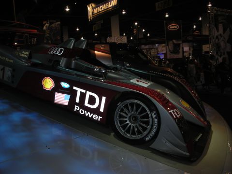 1998 Ford Crown Victoria - silver and dark-red Audi Le Mans prototype race car with Shell and TDI Power livery sits under spotlights at an expo.