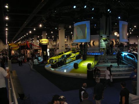 1998 Ford Crown Victoria - wide elevated view of a busy indoor automotive trade show floor with a central yellow show car, booths, and crowds.