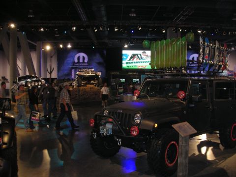 1998 Ford Crown Victoria - dark lifted Jeep Wrangler with snowboards on a roof rack stands at a Mopar indoor display with crowds and screens.