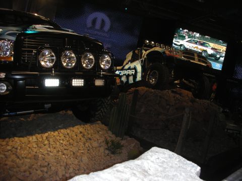 1998 Ford Crown Victoria - dark truck with a four-light bar and a Mopar-liveried off-road trophy truck climb a staged rock diorama under a video screen.