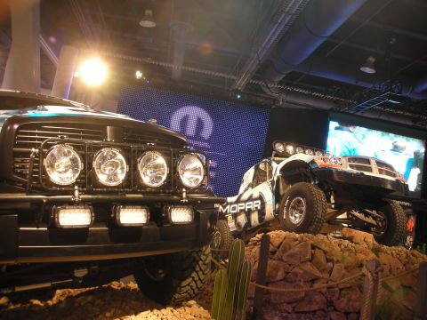 1998 Ford Crown Victoria - front-lit truck with stacked auxiliary lights and a white Mopar off-road trophy truck sit on a staged rock display under spotlights.