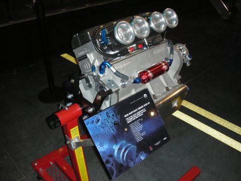 1998 Ford Crown Victoria - polished aluminum V8 race engine fitted with eight velocity stacks and blue lines sits on a red engine stand beside an info placard.