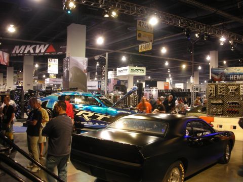 1998 Ford Crown Victoria - busy indoor auto show booth features a blue RT-badged car with raised hood and a matte-black muscle coupe among crowds.