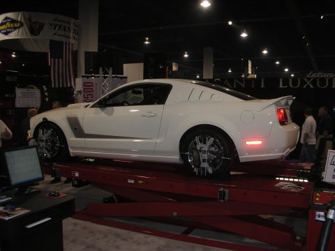 1998 Ford Crown Victoria - white two-door Mustang coupe with chrome wheels sits on a red chassis dyno rig at an indoor automotive show.