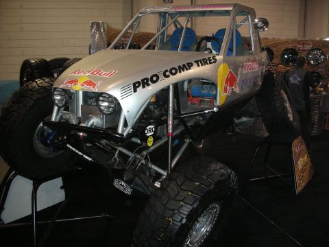 1998 Ford Crown Victoria - silver tube-chassis off-road rock buggy with Red Bull and Pro Comp Tires livery sits on large mud tires at an indoor show.