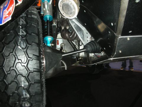 1998 Ford Crown Victoria - Underbody detail of an off-road buggy showing a blue coilover shock, CV axle, and chunky tire at an indoor display.