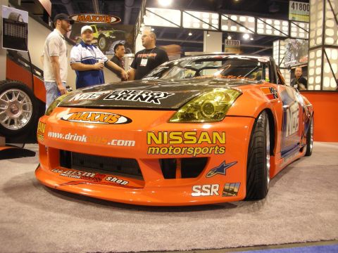 1998 Ford Crown Victoria - Side profile of an orange Nissan 350Z drift car with NOS, Maxxis and SSR graphics, surrounded by people at a trade show.