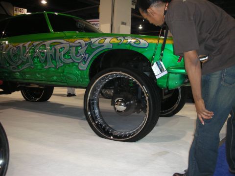 1998 Ford Crown Victoria - man bends to inspect a green graffiti-airbrushed SUV sitting on oversized black-and-chrome show wheels.