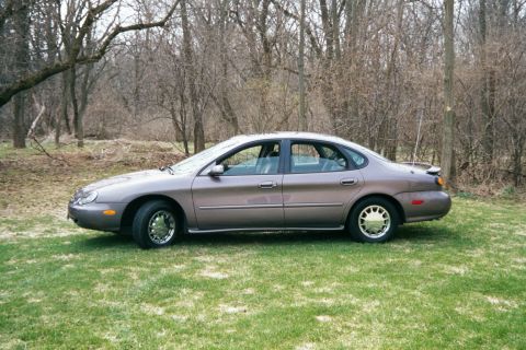 1996 Ford Taurus - This 1996 Ford Taurus is seen from the side, parked in a grassy area with trees behind it.